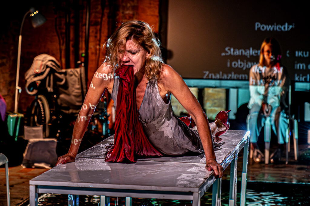 Seria zdjęć z eksperymentalnego spektaklu performatywnego rozgrywającego się na podłodze pokrytej cienką warstwą wody, z publicznością siedzącą bardzo blisko sceny. Na zdjęciach widać aktorkę wykonującą intensywne, fizyczne działania — chodzenie boso po wodzie, wspinanie się na metalowe stoły, przeciąganie materiałów, zanurzanie rąk i ciała w wodzie. Na ekranie za nią wyświetlane są fragmenty tekstów, które odbijają się także na jej ciele. Światło jest teatralne i kontrastowe, a przestrzeń przypomina surową halę z ceglaną ścianą, metalowymi konstrukcjami i widocznym sprzętem scenicznym. Publiczność obserwuje występ z bliska, otaczając scenę z dwóch stron.