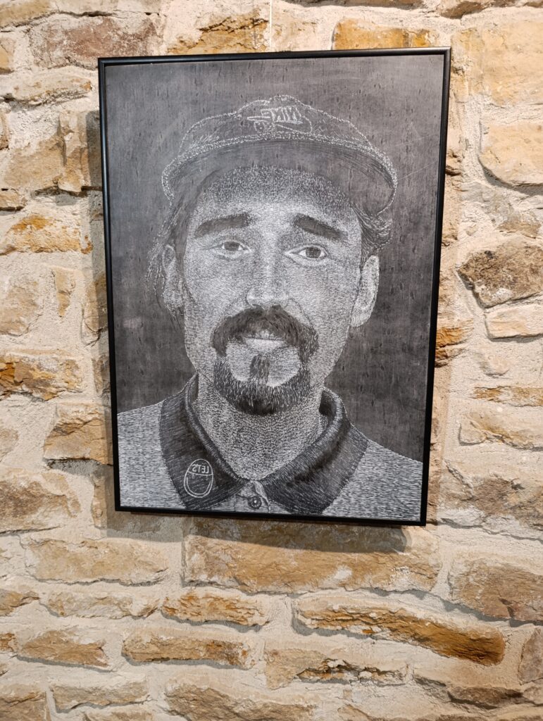 Oprawiony czarno-biały linoryt przedstawiający frontalny portret mężczyzny w czapce, z wąsami i brodą. Grafika zawieszona na kamiennej ścianie w przestrzeni wystawowej, z wyraźnie widoczną fakturą gęstych nacięć budujących światłocień twarzy.