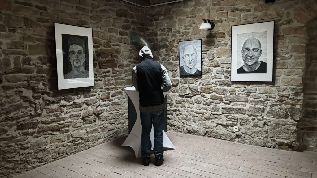 Wnętrze kameralnej przestrzeni wystawowej z kamiennymi ścianami. Na ścianach wiszą oprawione czarno-białe portrety wykonane w technice linorytu. Pośrodku sali stoi osoba tyłem do obiektywu, oglądająca prace przy wysokim stojącym stoliku. Scena oświetlona punktowym światłem ściennym.