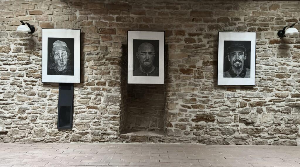 Fragment ekspozycji w kamiennej przestrzeni wystawowej. Na ścianie zawieszone są trzy oprawione czarno-białe linoryty przedstawiające męskie portrety, rozmieszczone równomiernie w jednej linii. Każda praca oświetlona jest punktową lampą ścienną, podkreślającą fakturę graficznych nacięć.