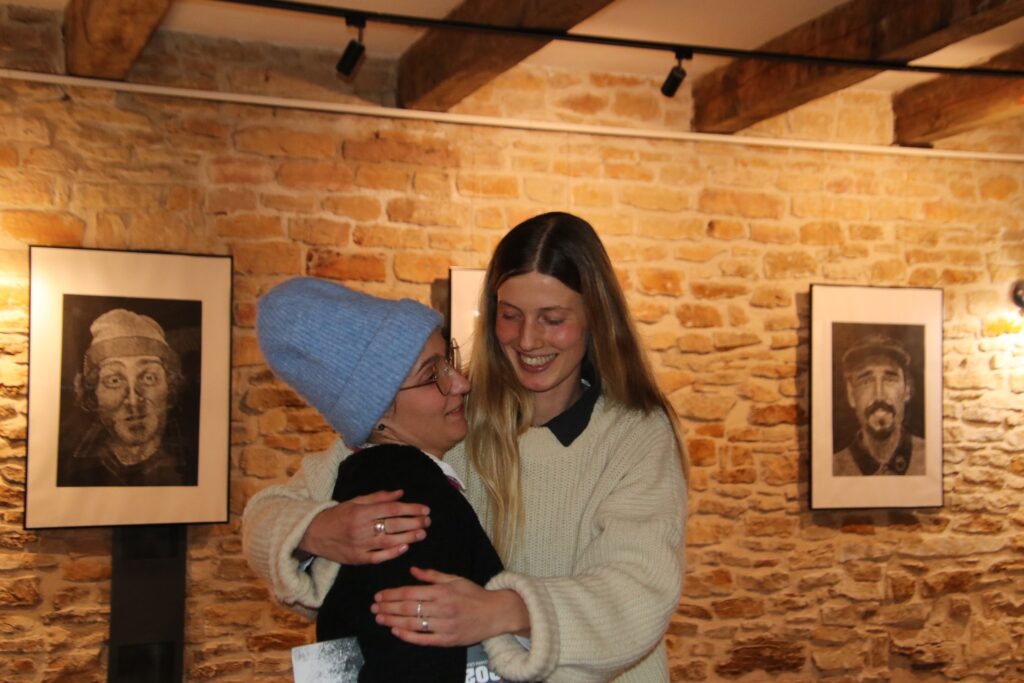 Dwie osoby obejmują się w przestrzeni wystawowej z kamiennymi ścianami. Stoją przed oprawionymi czarno-białymi linorytami przedstawiającymi męskie portrety. Scena oświetlona ciepłym światłem, oddająca atmosferę spotkania podczas wystawy.