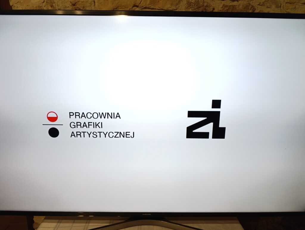 Biała plansza z logotypem „Pracownia Grafiki Artystycznej” po lewej stronie, składającym się z nazwy i prostych symboli graficznych w kolorach czerwonym i czarnym. Po prawej stronie znajduje się duży, czarny znak typograficzny. Plansza wyświetlana na ekranie.