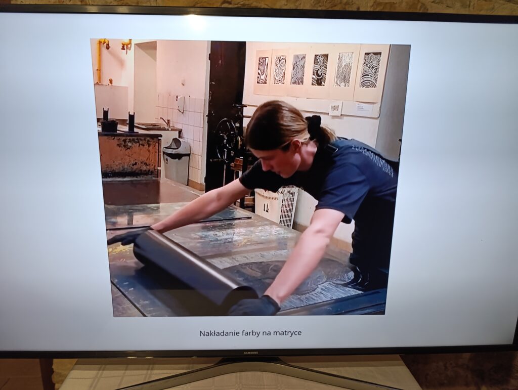 Osoba pracująca w pracowni graficznej nakłada farbę na matrycę linorytniczą za pomocą wałka. Pochylona nad stołem roboczym, w rękawiczkach ochronnych, wykonuje równomierne ruchy. W tle widoczne elementy wyposażenia pracowni oraz przykładowe grafiki na ścianie. Pod zdjęciem podpis: „Nakładanie farby na matrycę”.