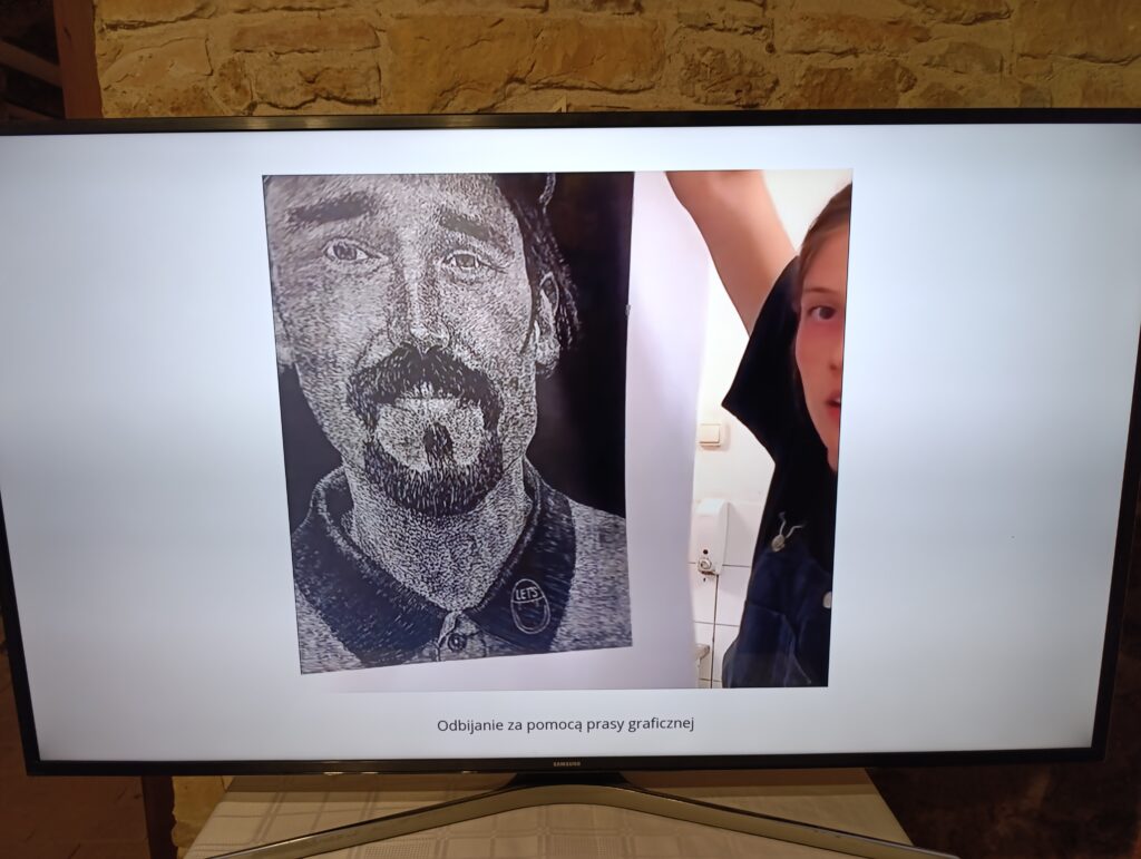 Czarno-biała odbitka linorytu przedstawiająca męski portret z wąsami i brodą, widoczna w trakcie procesu drukowania. Po prawej stronie kadru częściowo widoczna osoba obsługująca prasę graficzną, unosząca rękę. Pod zdjęciem podpis: „Odbijanie za pomocą prasy graficznej”.