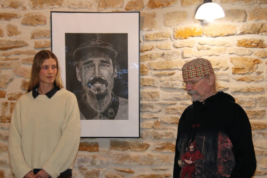 Dwie osoby stoją przed kamienną ścianą w przestrzeni wystawowej. Między nimi zawieszona jest oprawiona czarno-biała grafika — linoryt przedstawiający męski portret w czapce, z wąsami i brodą. Osoba po lewej stoi frontalnie, osoba po prawej spogląda w dół. Scena oświetlona lampą ścienną nad grafiką.