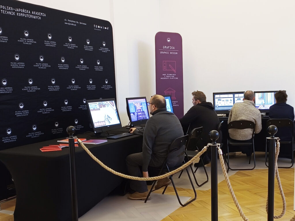 Stanowisko z edukacyjnymi grami przygotowanymi przez studentów PJATK, prezentowane w ramach współpracy z PAN Muzeum Ziemi i Muzeum Ewolucji PAN. Kilka osób siedzi przy komputerach i testuje interaktywne gry o tematyce przyrodniczej. Po lewej stronie znajduje się czarne tło z logo PJATK, po prawej — banner kierunku Grafika. Przestrzeń jest odgrodzona słupkami z linami.