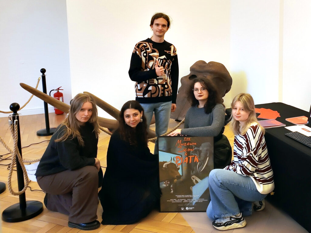 Grupa pięciu studentów PJATK pozujących przy instalacji w PAN Muzeum Ziemi. Cztery osoby kucają, trzymając plakat projektu „PAN Muzeum Ziemi & PJATK”, a jedna stoi za nimi. W tle widoczne są elementy ekspozycji muzealnej, m.in. duże skamieniałości, oraz stół z materiałami promocyjnymi PJATK.