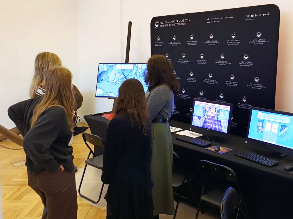 Grupa studentek PJATK ogląda edukacyjne gry na monitorach podczas prezentacji projektu w PAN Muzeum Ziemi. Na biurkach stoją komputery z uruchomionymi interaktywnymi aplikacjami, a w tle widoczna jest czarna ścianka z logo Polsko-Japońskiej Akademii Technik Komputerowych. Studentki stoją przy stanowisku i rozmawiają o wyświetlanych grach.