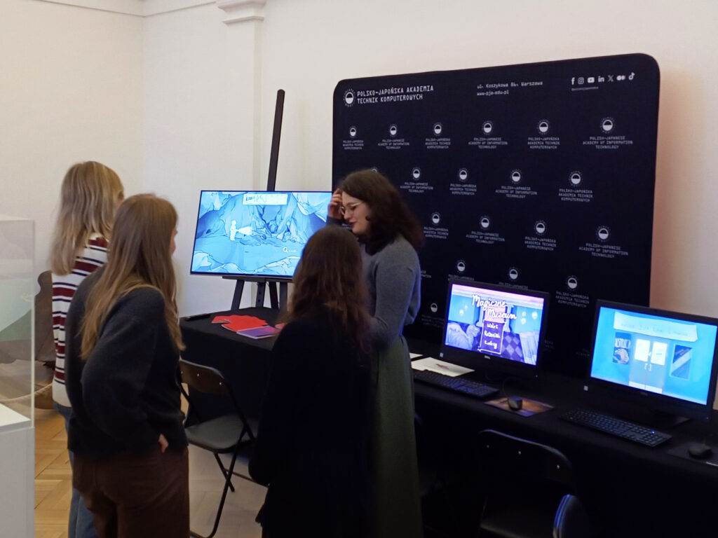 Studentki PJATK stoją przy stanowisku z edukacyjnymi grami prezentowanymi w PAN Muzeum Ziemi. Na monitorach widać interaktywne aplikacje stworzone przez studentów, a w tle znajduje się czarna ścianka z logo Polsko-Japońskiej Akademii Technik Komputerowych. Jedna z uczestniczek tłumaczy działanie gry pozostałym.