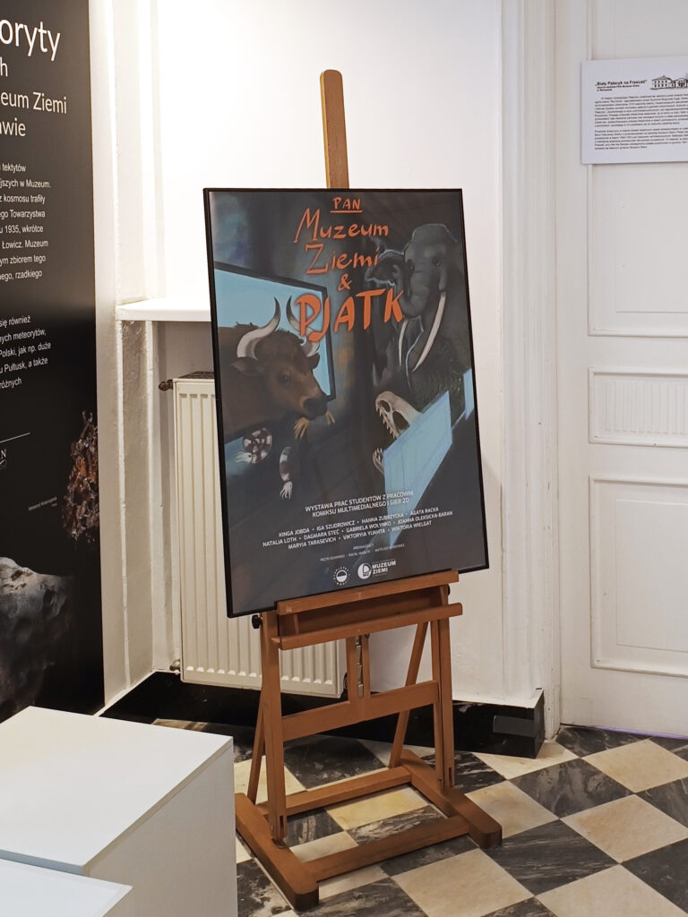 Plakat wystawy „PAN Muzeum Ziemi & PJATK” ustawiony na drewnianej sztaludze w przestrzeni muzealnej. Ilustracja przedstawia zwierzęta prehistoryczne, m.in. tura i mamuta. W tle widoczne są fragmenty ekspozycji i tablice informacyjne muzeum, a na podłodze charakterystyczna czarno-biała posadzka.
