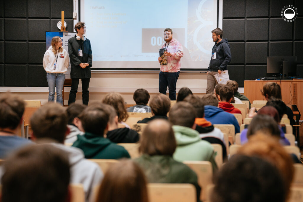 Czworo prowadzących stoi przed uczestnikami w sali wykładowej podczas Game Jam 2025 na PJAIT. Za nimi widoczny jest duży ekran z prezentacją dotyczącą przedstawiania zespołów. Publiczność siedzi w rzędach drewnianych krzeseł, skierowana w stronę sceny. Po prawej stronie znajduje się biurko z komputerem, a w rogu ekranu widnieje logo PJAIT.