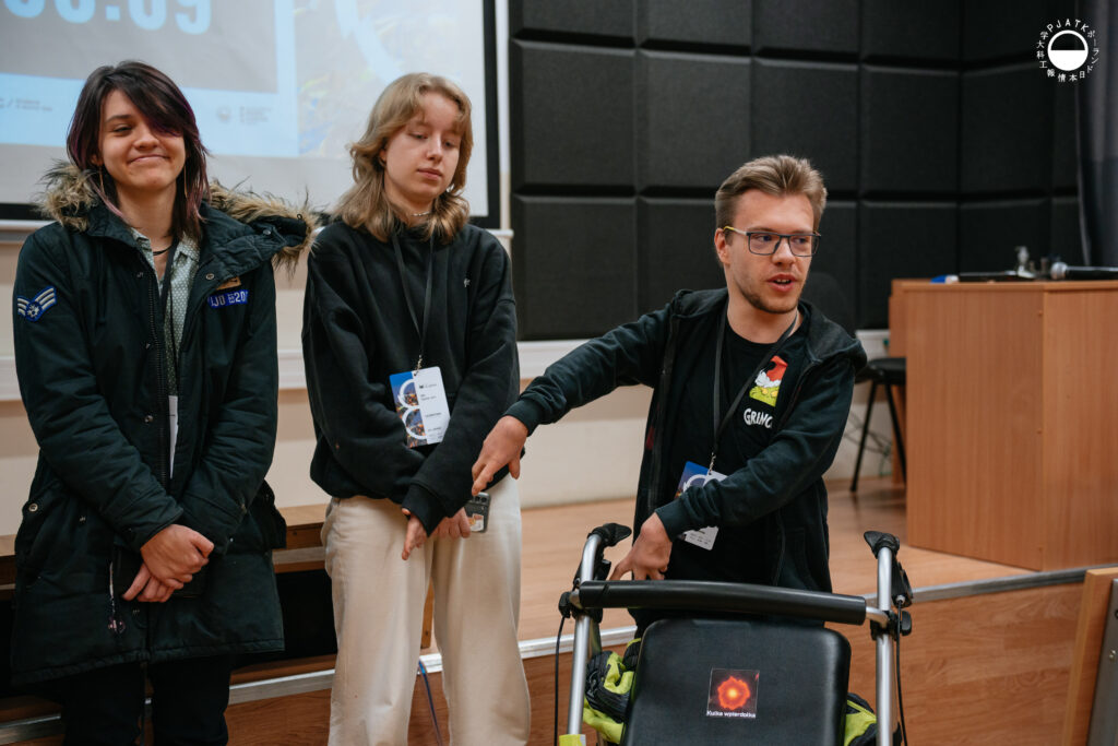 Troje uczestników Game Jamu 2025 stoi przed publicznością w sali PJAIT. Dwie osoby stoją obok siebie, a trzecia – mężczyzna z chodzikiem – przedstawia się, gestykulując rękami. Wszyscy mają identyfikatory wydarzenia na smyczach. W tle widoczny jest fragment ekranu z prezentacją oraz czarne panele akustyczne sali.