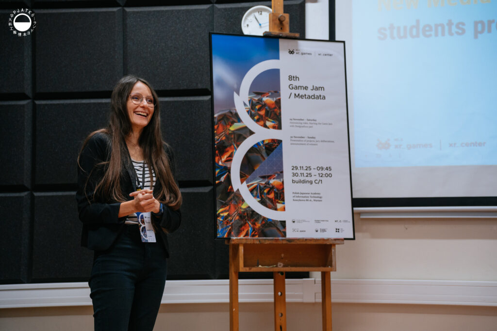 Uśmiechnięta prowadząca stoi na tle czarnych paneli akustycznych w sali PJAIT podczas Game Jamu 2025. Obok niej znajduje się duży plakat 8th Game Jam / Metadata z programem wydarzenia i datami 29–30.11.2025. Po prawej stronie widoczny jest fragment ekranu z prezentacją.