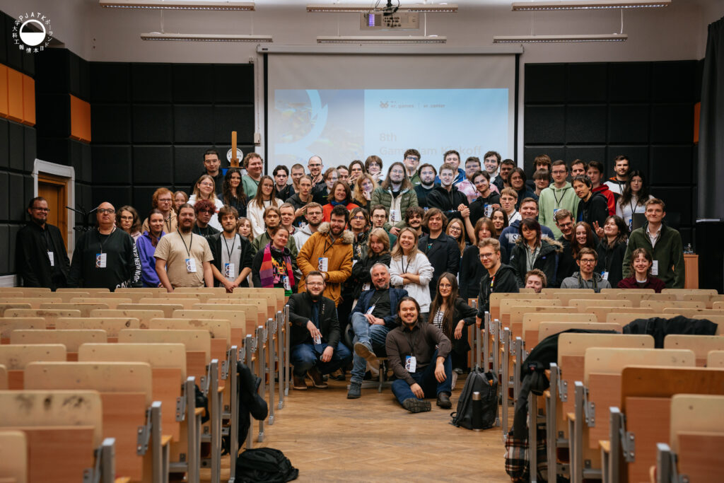 Duża grupa uczestników 8th Game Jam / Metadata na PJAIT pozuje do wspólnego zdjęcia w sali wykładowej. Ludzie stoją i siedzą w kilku rzędach między pustymi ławkami, uśmiechając się w stronę aparatu. W tle widać ekran z prezentacją wydarzenia oraz czarne panele akustyczne sali.
