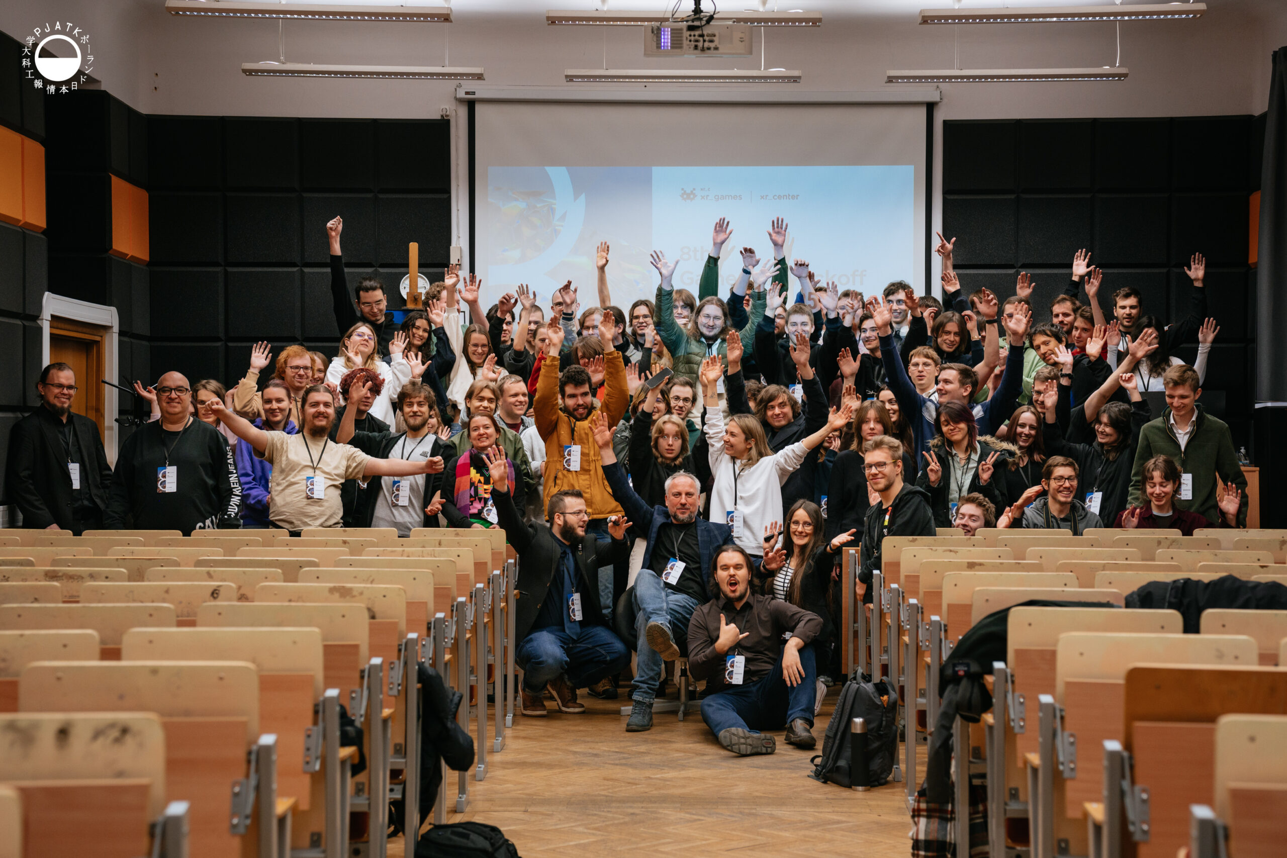 Duża grupa uczestników 8th Game Jam / Metadata pozuje w sali wykładowej PJAIT, radośnie unosząc ręce do góry. Uczestnicy stoją i siedzą między rzędami pustych ławek, uśmiechają się i wykonują energiczne gesty. W tle widoczny jest ekran z prezentacją wydarzenia oraz czarne panele akustyczne.