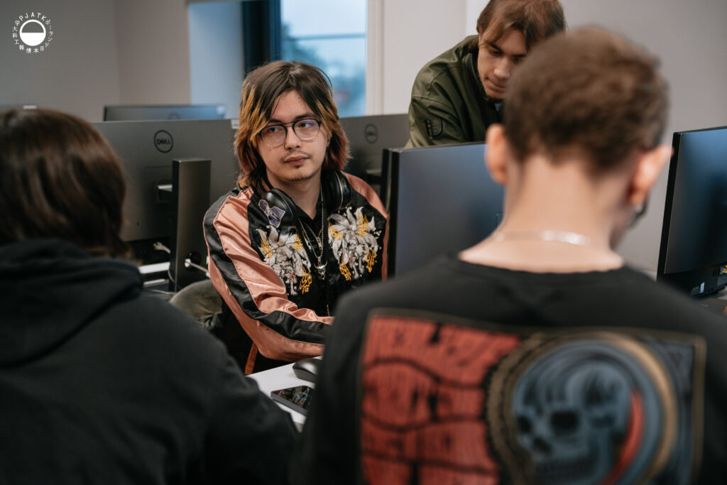 Grupa uczestników Game Jamu pracuje przy stanowiskach komputerowych w laboratorium PJAIT. Na pierwszym planie widać młodego mężczyznę w różowo-czarnej kurtce z kwiatowym wzorem, z założonymi na szyi słuchawkami, patrzącego w stronę rozmówcy. Wokół niego pozostali członkowie zespołu pochylają się nad monitorami.