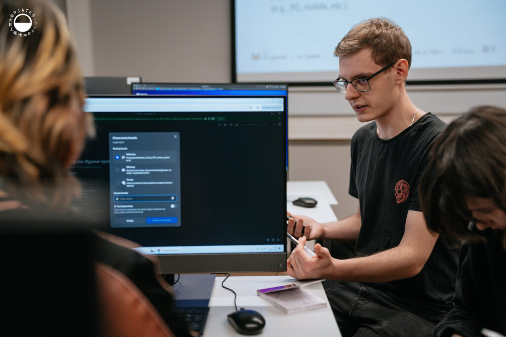 Uczestnik Game Jamu siedzi przy stanowisku komputerowym w laboratorium PJAIT, tłumacząc coś członkom swojego zespołu. Na monitorze przed nim widoczne jest okno ustawień w aplikacji. Obok niego druga osoba pochyla się nad własnym ekranem, a na biurku leżą notatki i myszka.