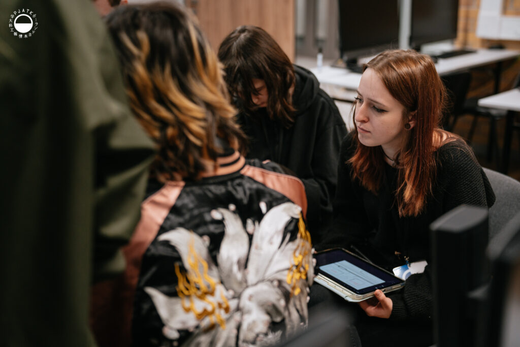 Uczestniczka Game Jamu siedzi z tabletem na kolanach i uważnie słucha rozmowy zespołu w laboratorium PJAIT. Obok niej dwie osoby pochylają się nad swoimi notatkami i ekranami, a na pierwszym planie widać fragment kurtki jednego z członków grupy.