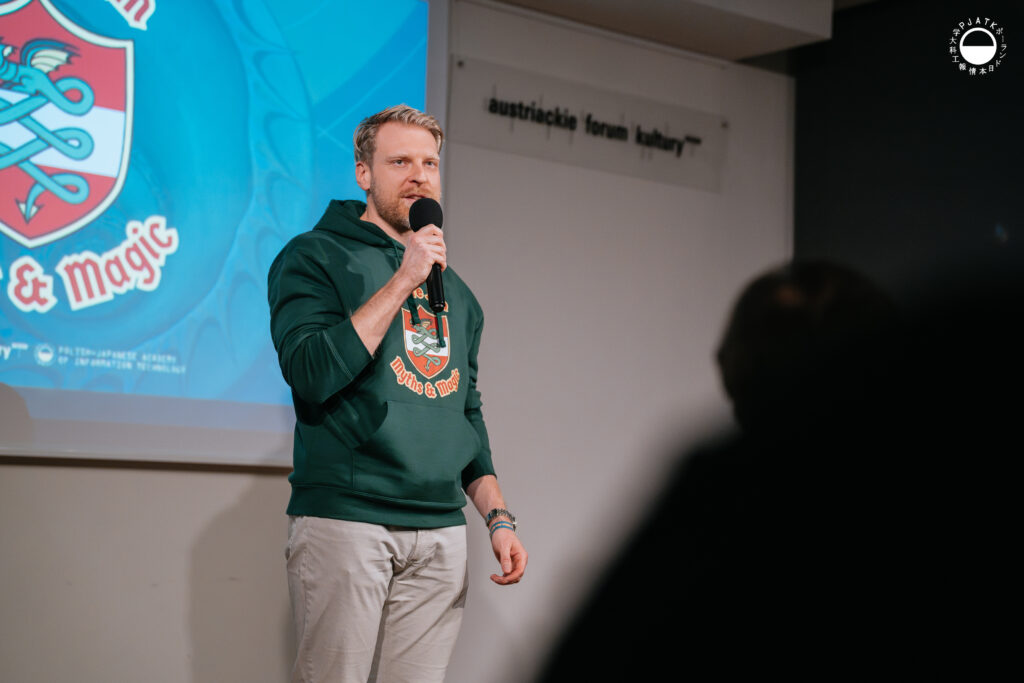 Mężczyzna w zielonej bluzie Culture Jam: Myths & Magic przemawia do publiczności na scenie Austriackiego Forum Kultury, trzymając mikrofon.