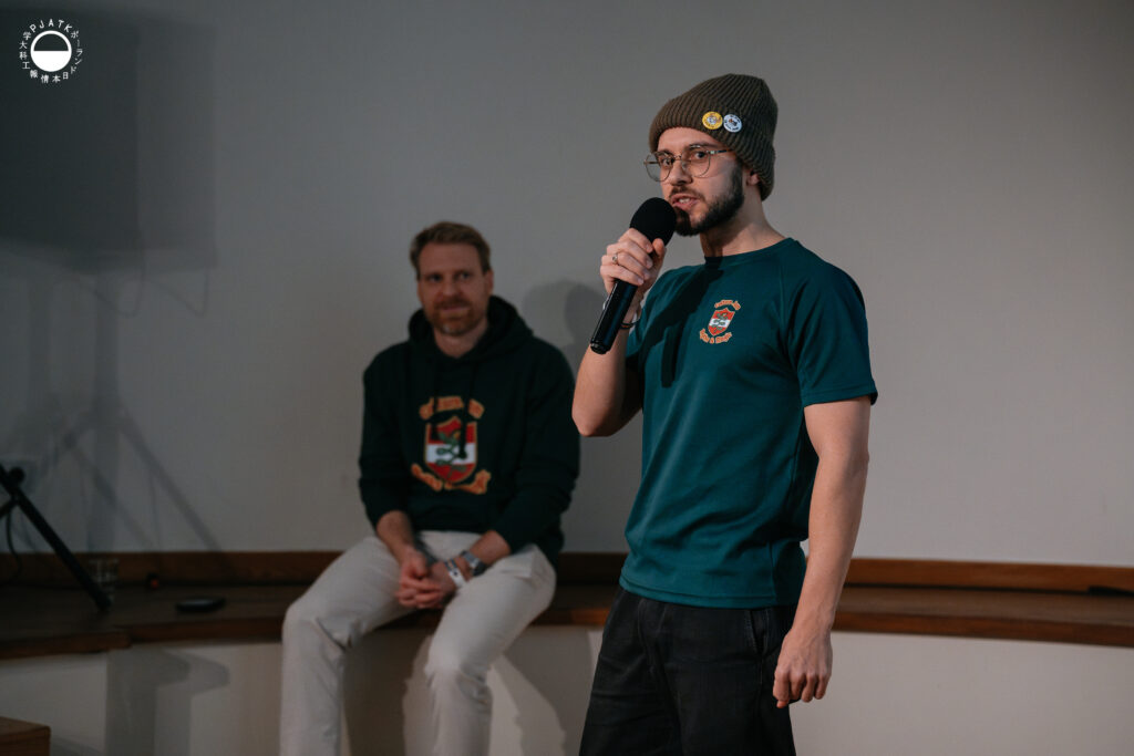 Mężczyzna w czapce i koszulce Culture Jam przemawia do mikrofonu, a za nim na ławce siedzi drugi uczestnik wydarzenia.