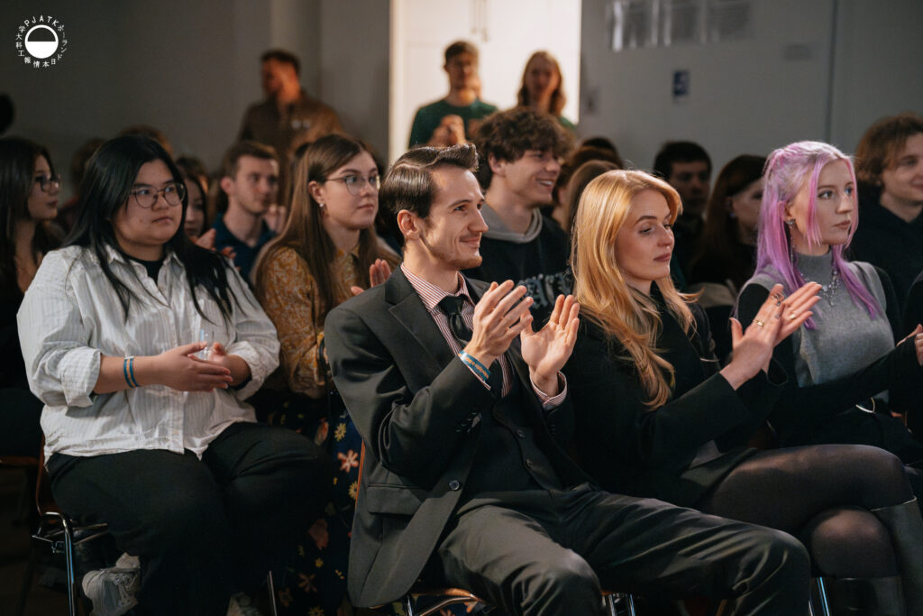 Publiczność wydarzenia Culture Jam siedzi w rzędach i bije brawo, uśmiechając się w stronę sceny.
