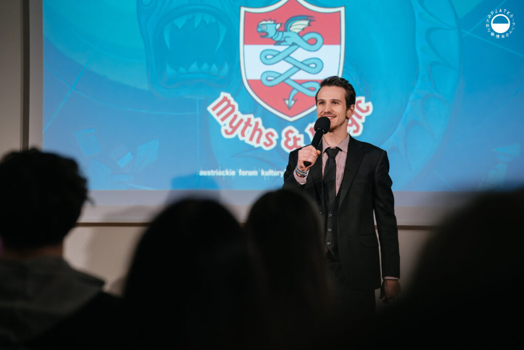 Prowadzący w garniturze przemawia do mikrofonu na tle slajdu Culture Jam: Myths & Magic.