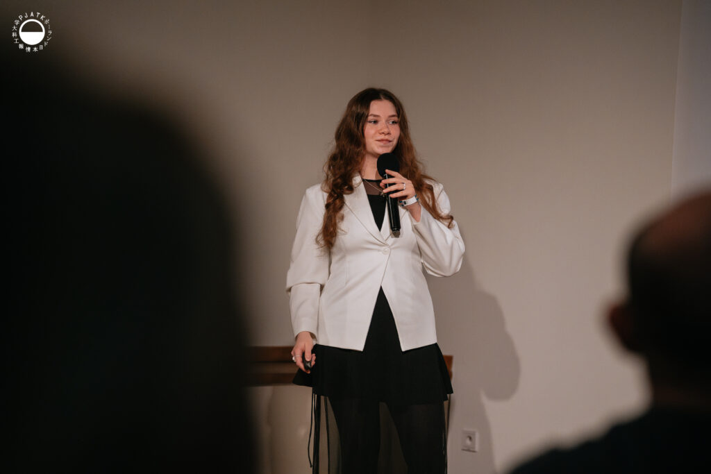 Kobieta w białej marynarce przemawia do mikrofonu na scenie podczas prezentacji.