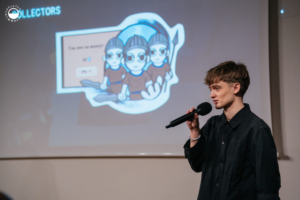 Prezentujący trzyma mikrofon i opowiada o grze, stojąc obok slajdu z ilustracją trzech kreskówkowych postaci.