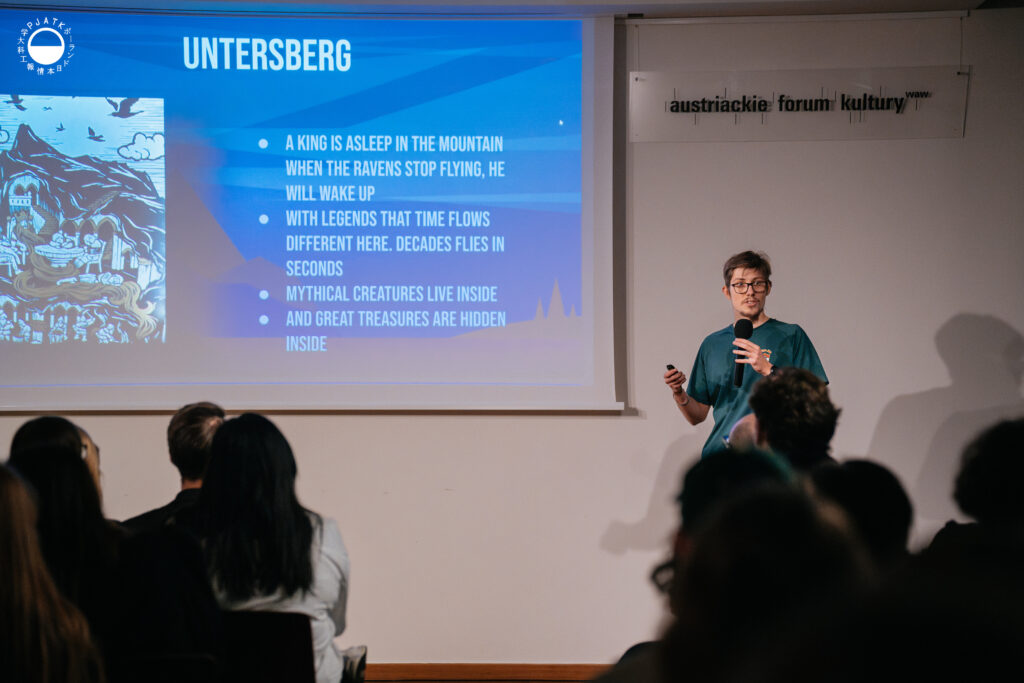 Prezentująca osoba omawia projekt „Untersberg”, stojąc z mikrofonem obok slajdu z opisem legendy i ilustracją góry.