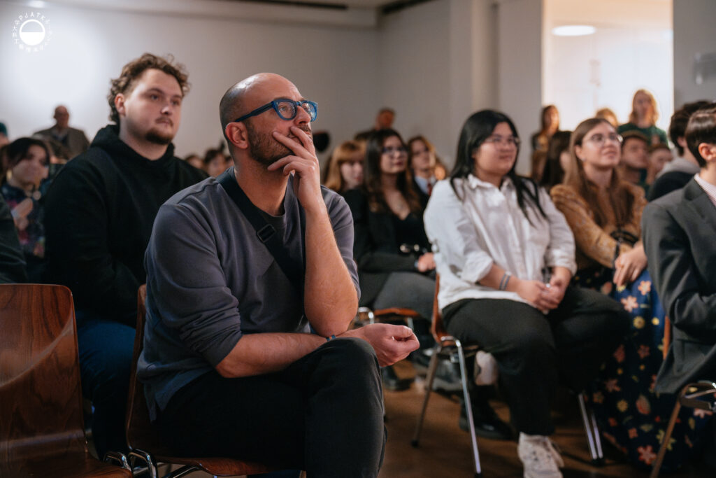 Publiczność uważnie słucha prezentacji; mężczyzna na pierwszym planie siedzi z ręką przy twarzy, a za nim widać skupionych uczestników wydarzenia.