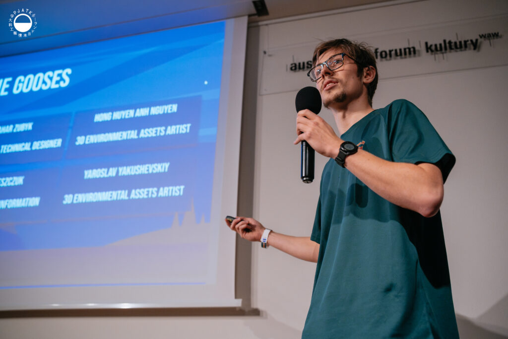 Prezentujący w zielonej koszulce omawia skład zespołu, stojąc z mikrofonem obok slajdu z listą nazwisk.