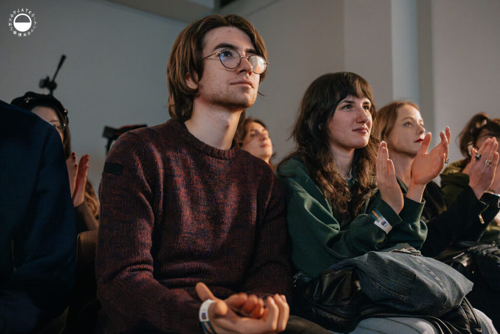 Uczestnicy wydarzenia siedzą w pierwszym rzędzie i biją brawo, uważnie patrząc w stronę sceny.