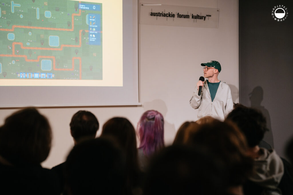 Prelegent w jasnej bluzie i czapce prezentuje fragment gry wyświetlony na ekranie, zwrócony w stronę publiczności.