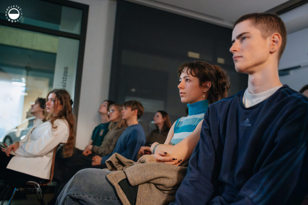 Grupa uczestników siedzi w sali i uważnie słucha prezentacji, patrząc w stronę sceny.