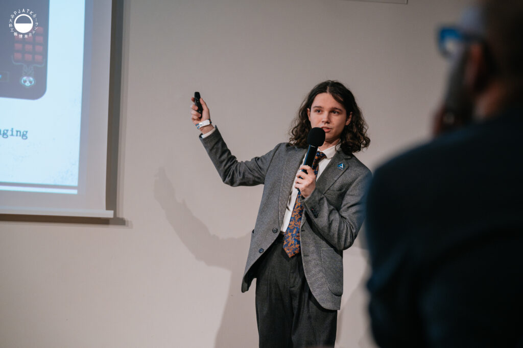 Prelegent w szarym garniturze prezentuje slajd, trzymając mikrofon i pilot, wskazując ręką w stronę ekranu.