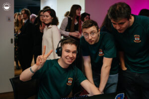 Trzech członków zespołu w zielonych koszulkach Culture Jam stoi przy komputerze; jeden z nich uśmiecha się do kamery i pokazuje znak pokoju.