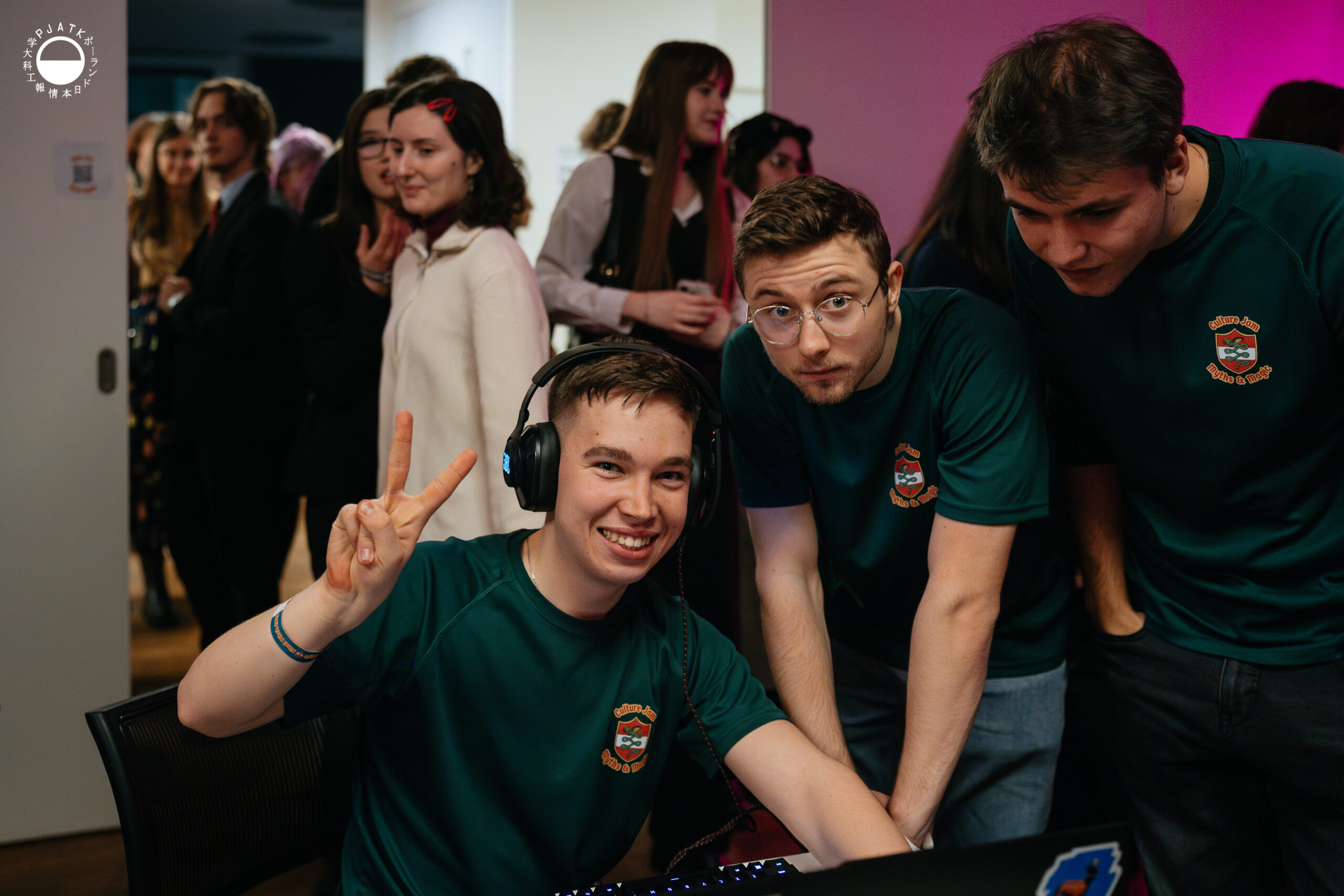 Trzech członków zespołu w zielonych koszulkach Culture Jam stoi przy komputerze; jeden z nich uśmiecha się do kamery i pokazuje znak pokoju.