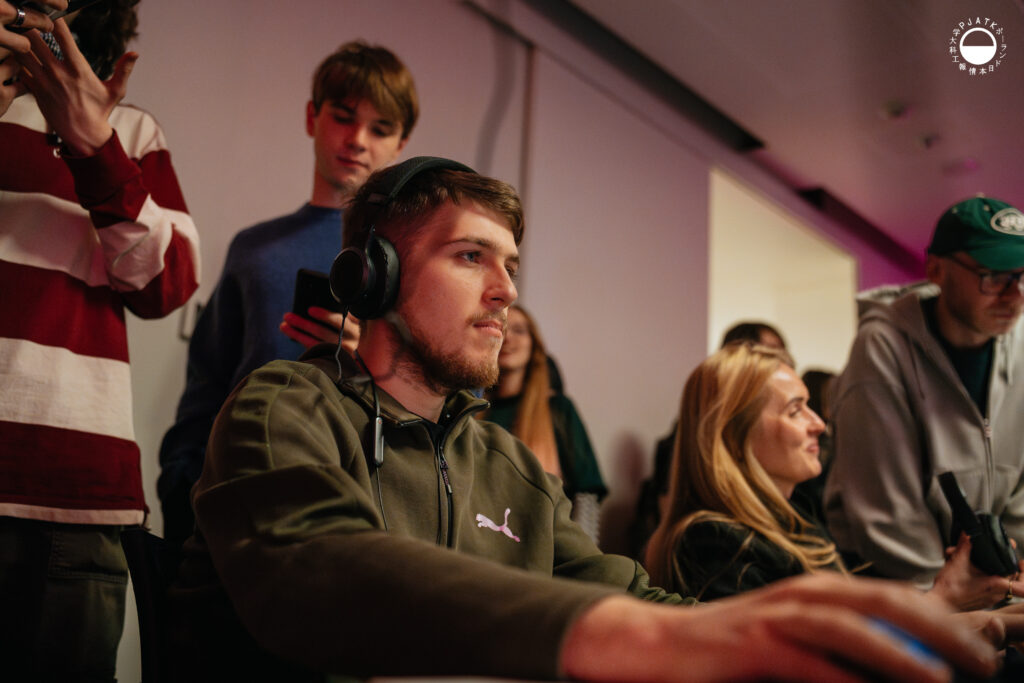 Uczestnik w słuchawkach gra na komputerze podczas prezentacji gier, otoczony obserwującymi go osobami.