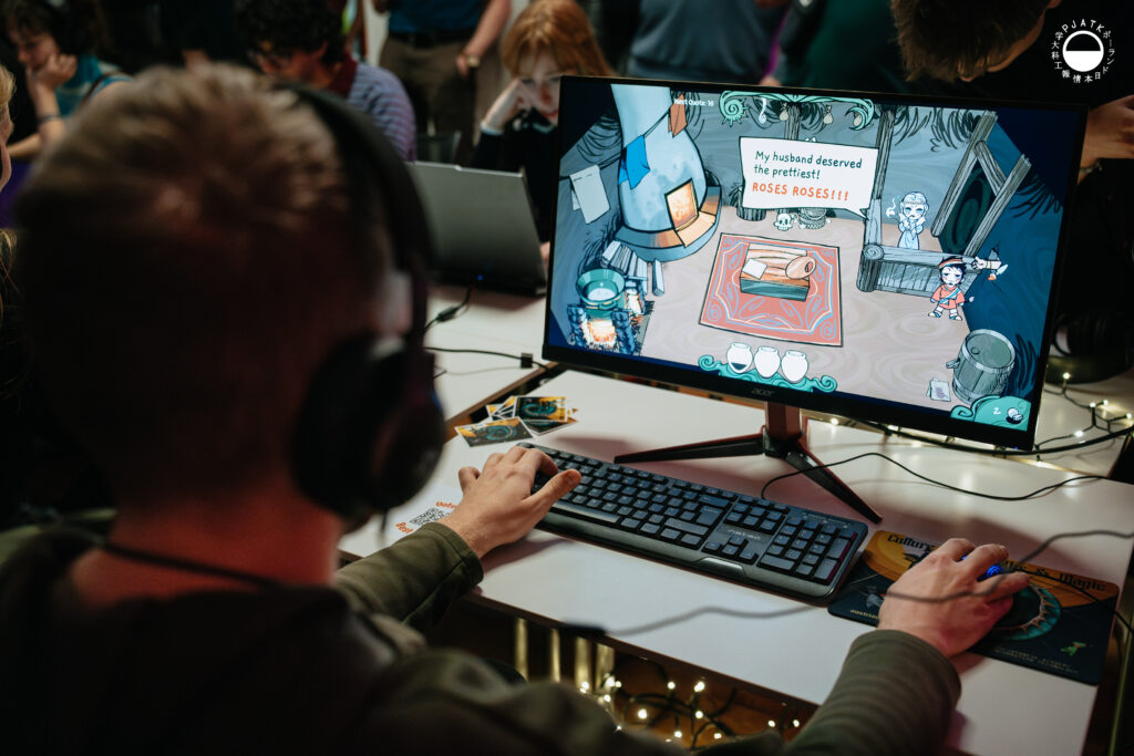 Osoba w słuchawkach gra na zaprezentowaną grę przy komputerze; na monitorze widać kreskówkową scenę z dialogiem „My husband deserved the prettiest! ROSES ROSES!!!”.