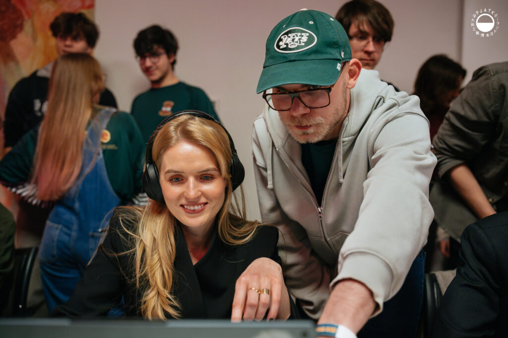 Uśmiechnięta uczestniczka w słuchawkach gra na komputerze, a stojący obok mężczyzna w czapce i okularach wskazuje jej coś na ekranie.