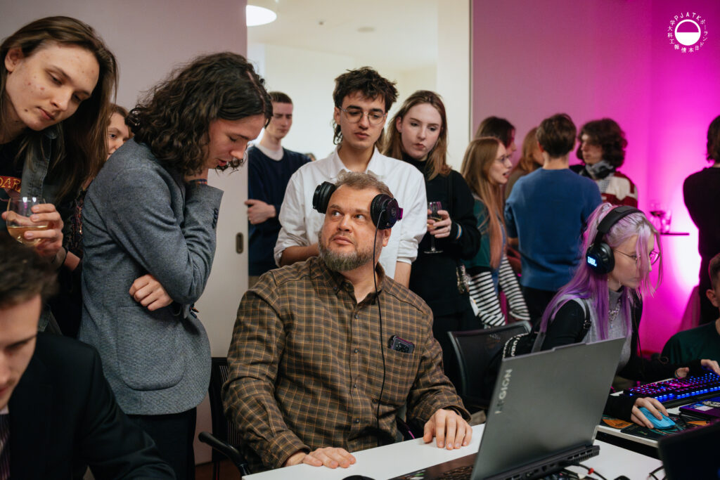 Grupa uczestników stoi wokół stanowiska z grami; mężczyzna w koszuli siedzi przy komputerze ze słuchawkami na szyi, patrząc na osobę obok, a obok niego druga uczestniczka gra na laptopie.