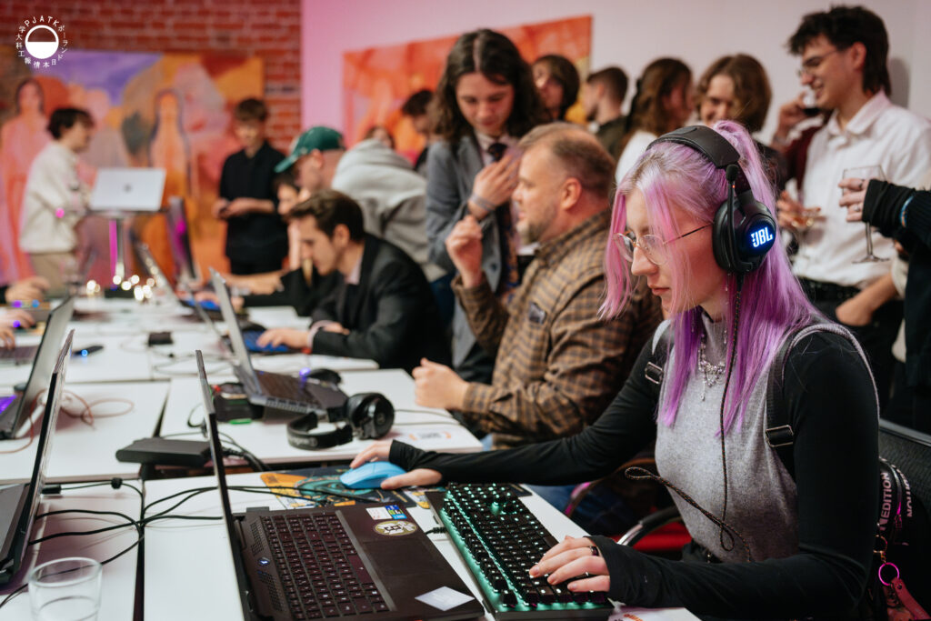 Uczestnicy testują gry przy długim stole z laptopami; na pierwszym planie osoba z fioletowymi włosami w słuchawkach gra na komputerze, a za nią inni rozmawiają i obserwują rozgrywkę.