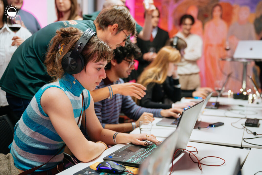 Uczestnicy w słuchawkach skupieni na grze przy laptopach; na pierwszym planie osoba w niebieskim swetrze gra, a prowadzący pochyla się, by udzielić wskazówki.