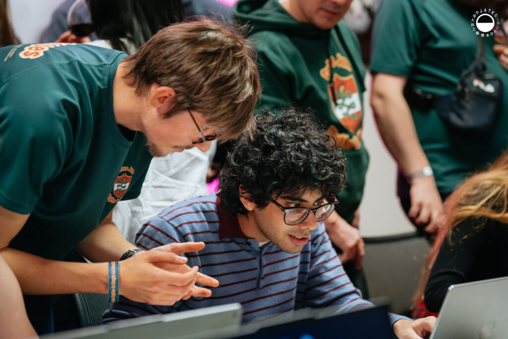 Uczestnik przy laptopie testuje grę, a członek zespołu w zielonej koszulce Culture Jam pochyla się obok, tłumacząc i gestykulując.