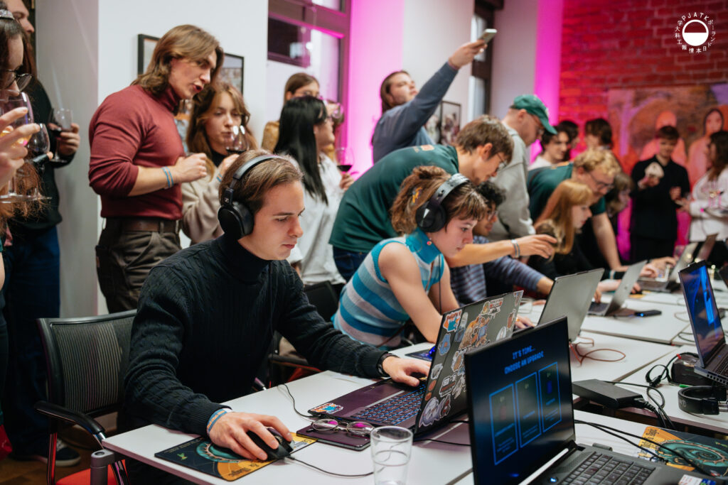 Uczestnicy w słuchawkach intensywnie grają przy laptopach, a za nimi tłum obserwuje rozgrywkę i robi zdjęcia podczas prezentacji gier.
