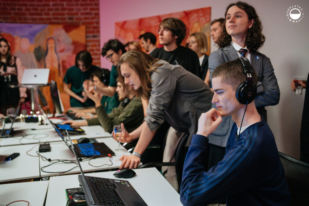 Uczestnicy grają przy laptopach, a za nimi tłum obserwuje i komentuje; na pierwszym planie gracz w słuchawkach skupia się na rozgrywce.