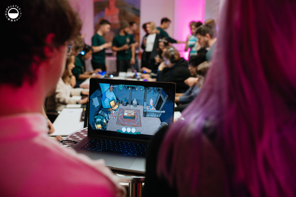 Uczestnicy oglądają laptop z uruchomioną grą; na ekranie widać kolorową, kreskówkową scenę, a w tle wiele osób testuje inne projekty.