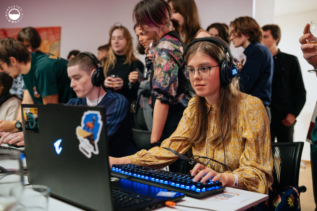 Uczestniczka w okrągłych okularach i słuchawkach gra przy laptopie, skupiona na rozgrywce, podczas gdy za jej plecami gromadzi się tłum obserwatorów.