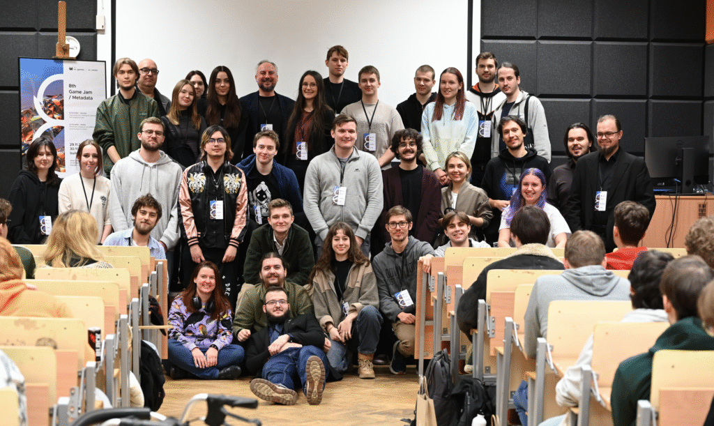 Grupa zwycięzców i uczestników Game Jamu 2025 pozuje do wspólnego zdjęcia w sali wykładowej PJAIT. Uczestnicy stoją i siedzą w kilku rzędach między ławkami, uśmiechając się w stronę aparatu. Po lewej stronie widoczny jest plakat wydarzenia ustawiony na sztaludze, a w tle ekran projekcyjny i czarne panele akustyczne.