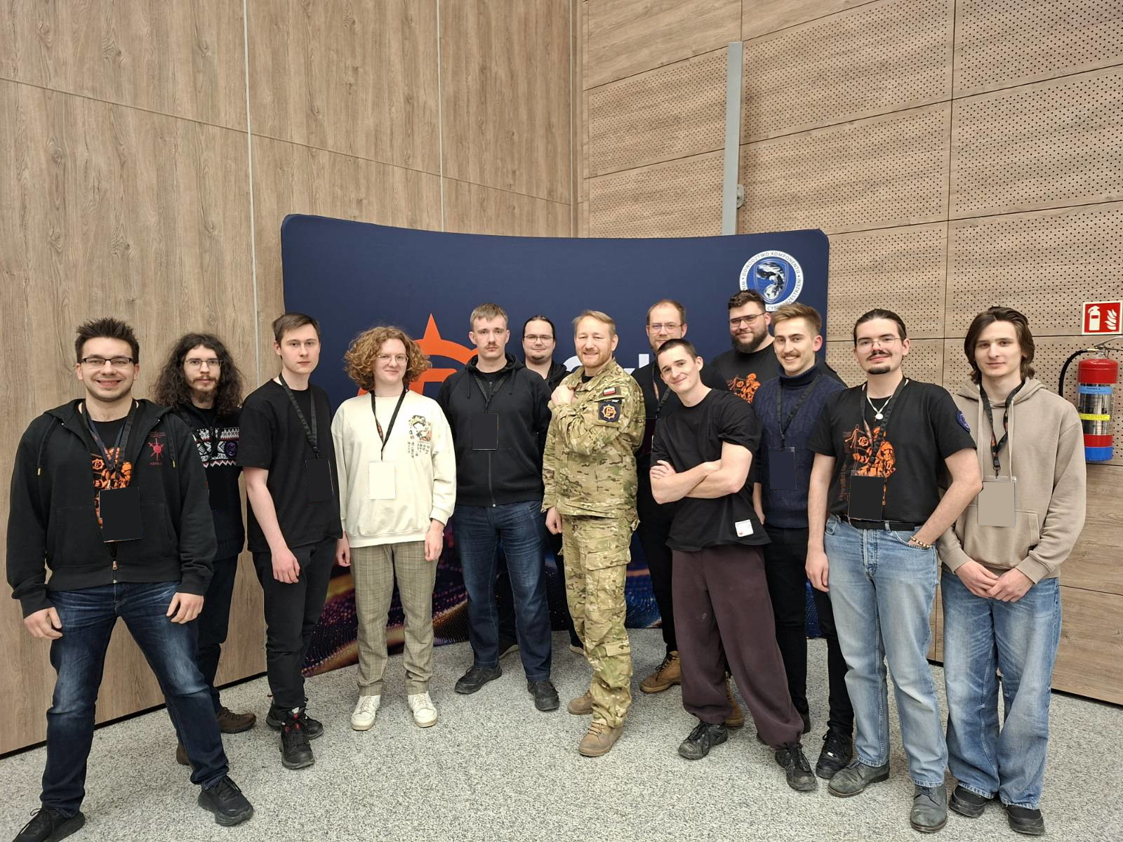 Grupowe zdjęcie 10 osób stojących w sali konferencyjnej przed dużą granatową ścianką z logo wydarzenia; w środku stoi osoba w mundurze wojskowym, pozostali uczestnicy są w casualowych ubraniach. W tle drewniane i perforowane panele ścienne oraz gaśnica po prawej.